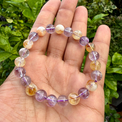 May include: A beaded bracelet featuring alternating round polished gemstones in shades of translucent purple amethyst and golden yellow citrine. The beads are strung on an elastic cord, creating a circular shape with a smooth, reflective surface.