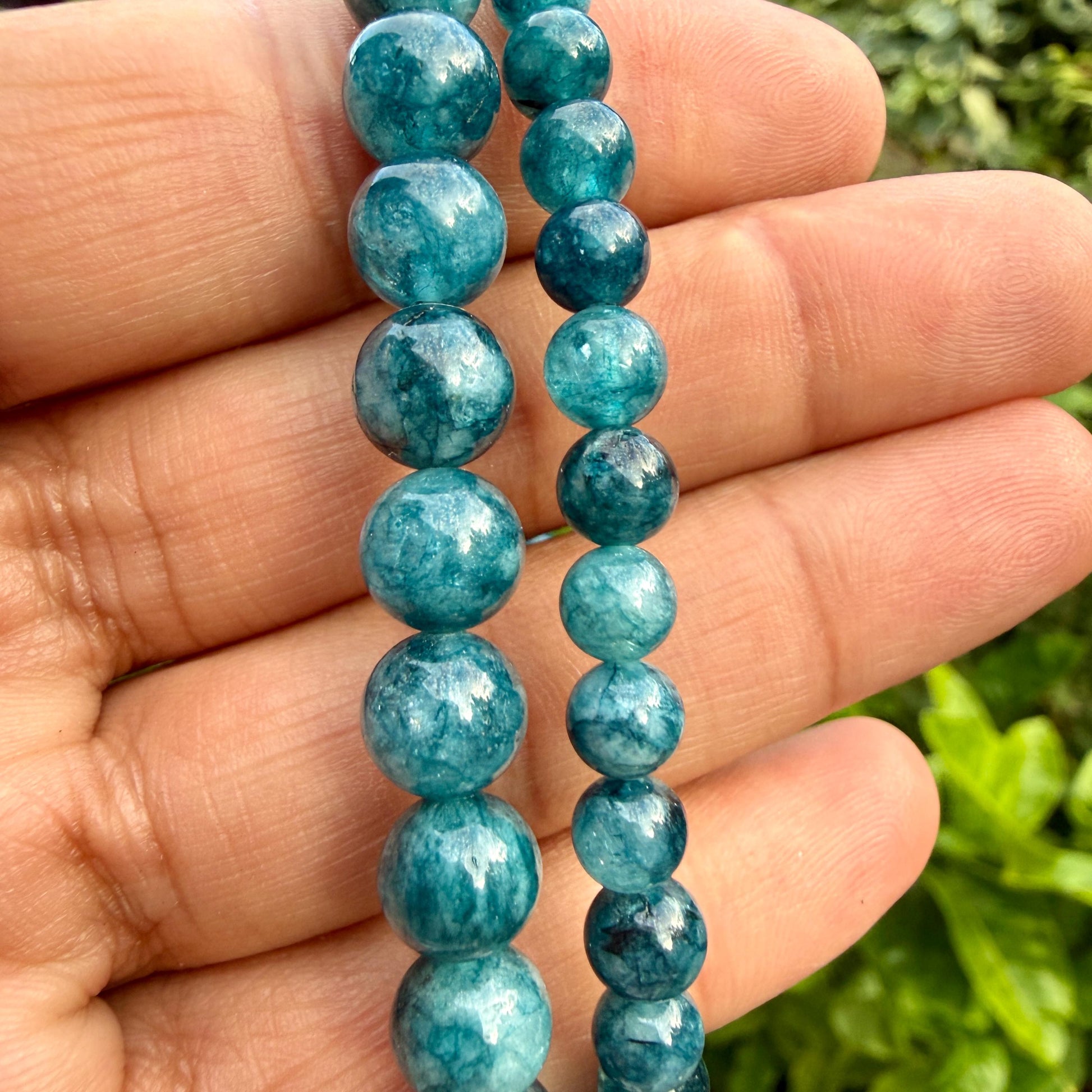 May include: Two strands of round, polished beads in varying shades of teal and deep blue. The beads feature a marbled, translucent texture with internal inclusions, arranged in two vertical rows of different sizes, showcasing a smooth finish.