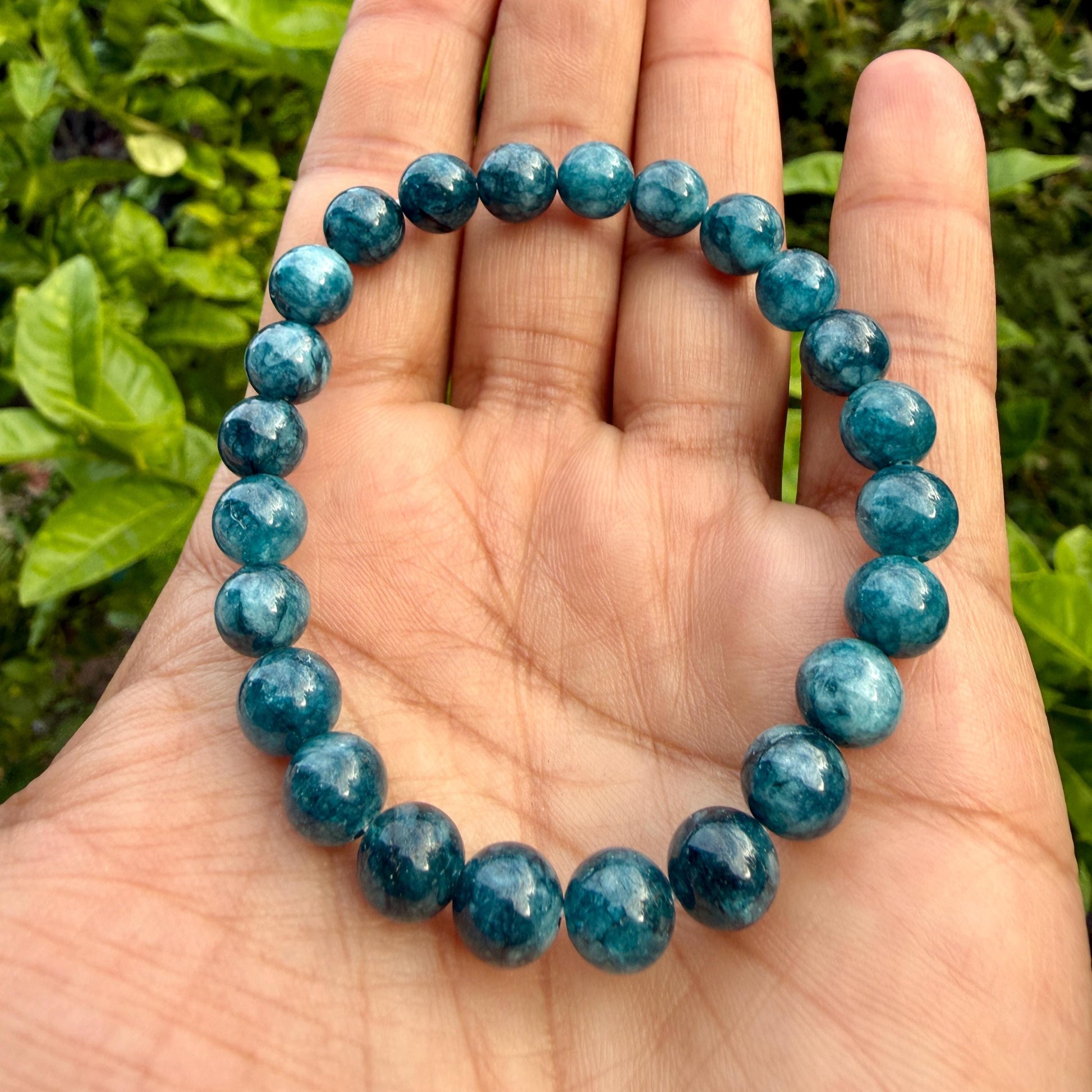 May include: A stretch bracelet composed of round, polished beads in varying shades of deep teal and blue. The beads exhibit natural marbling and translucent qualities, arranged in a continuous circular loop to form a single strand accessory.