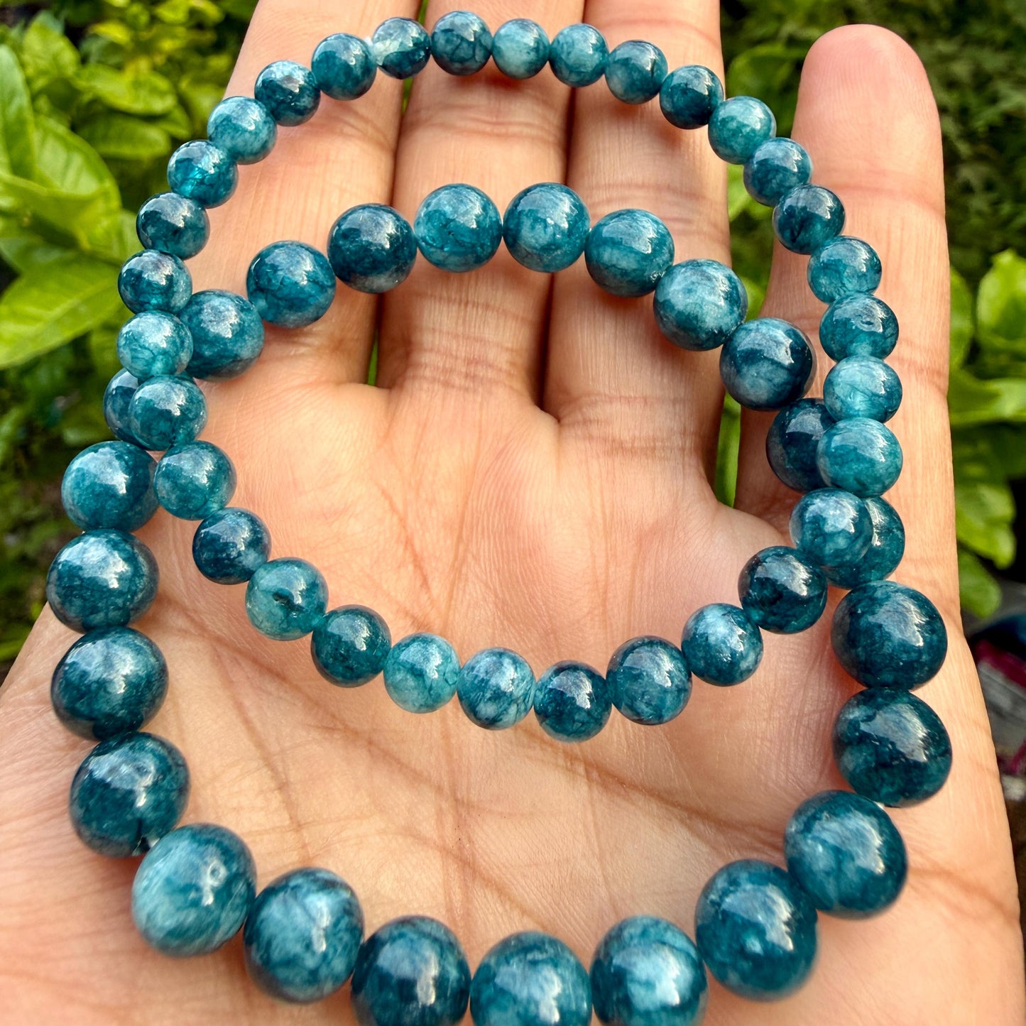 May include: A stretch bracelet composed of round, polished beads in a deep teal blue color. The beads feature natural internal marbling and variations in tone, creating a semi-translucent appearance throughout the circular strand of gemstone beads.