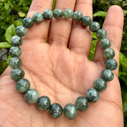 May include: A circular stretch bracelet composed of polished, round beads in varying shades of deep forest green and mossy grey. The beads feature a smooth, glossy finish with subtle internal marbling and natural inclusions throughout the stone.