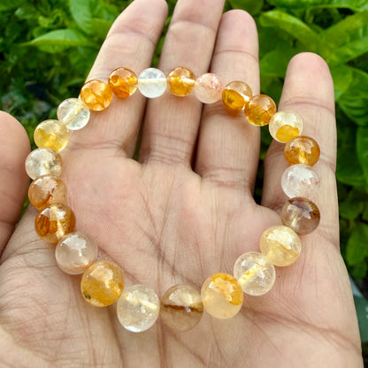May include: A stretch bracelet composed of round, polished beads in varying shades of translucent yellow, orange, and clear quartz. The beads exhibit natural internal inclusions and color gradients, arranged in a circular pattern on an elastic cord.